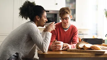 A couple shares a cup of coffee at their kitchen table. Sliced bread lays on the cutting board.